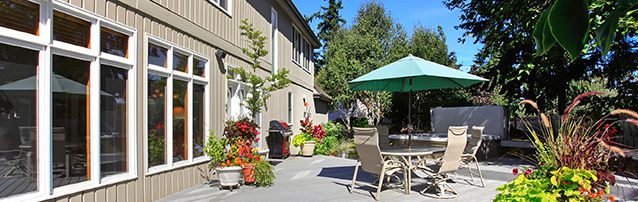 home patio in the sun, with dining table and chairs set with green umbrella. Next to a tan home with a row of large windows.