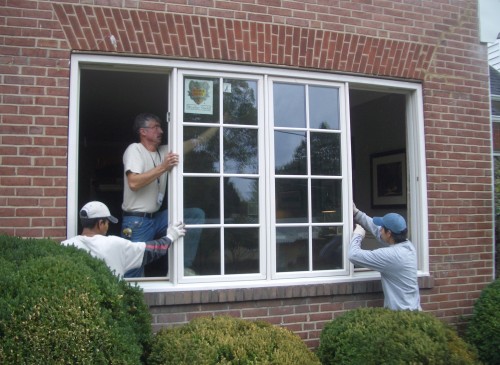 Team of window installers installing large white-frame window with grids in brick home.