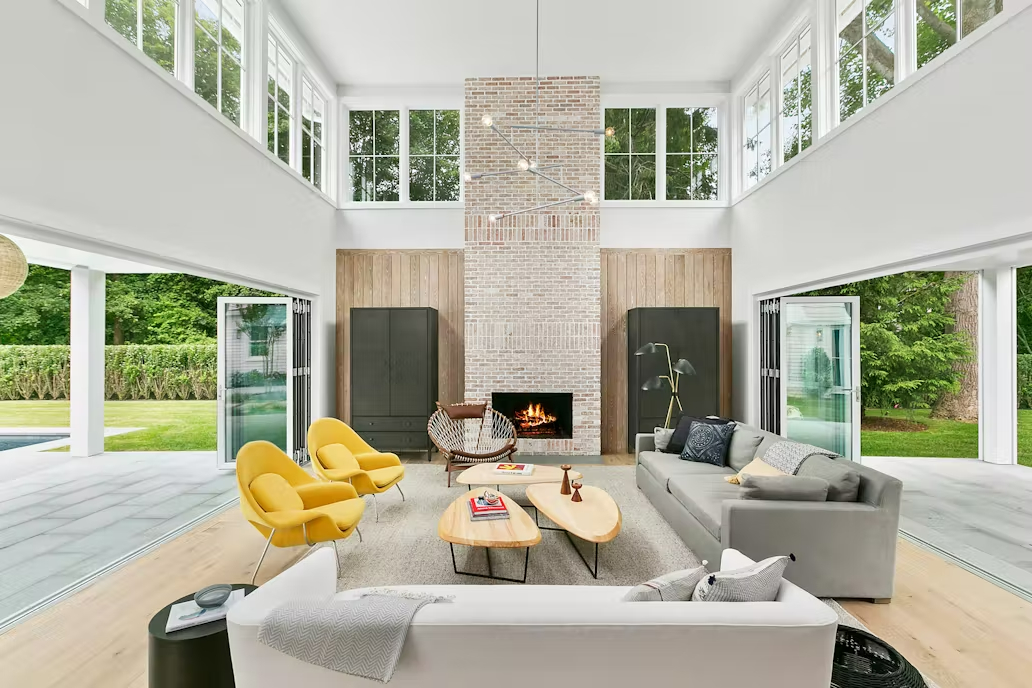 Large living room with high ceilings and large windows. Open accordion sliding glass doors on either side, leading to manicured lawn. Large two-story brick fireplace at far end of room. Light wood floors, gray couches and yellow chairs.