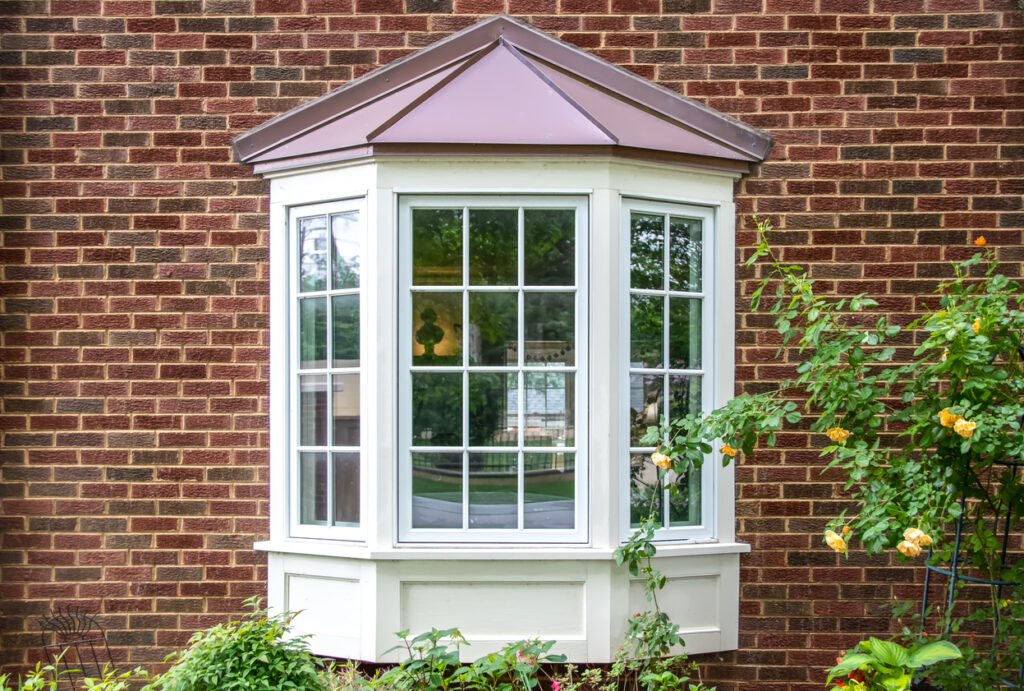 Bay window with copper roof on traditional brick home with flowers below and yellow roses extending out onto the window.